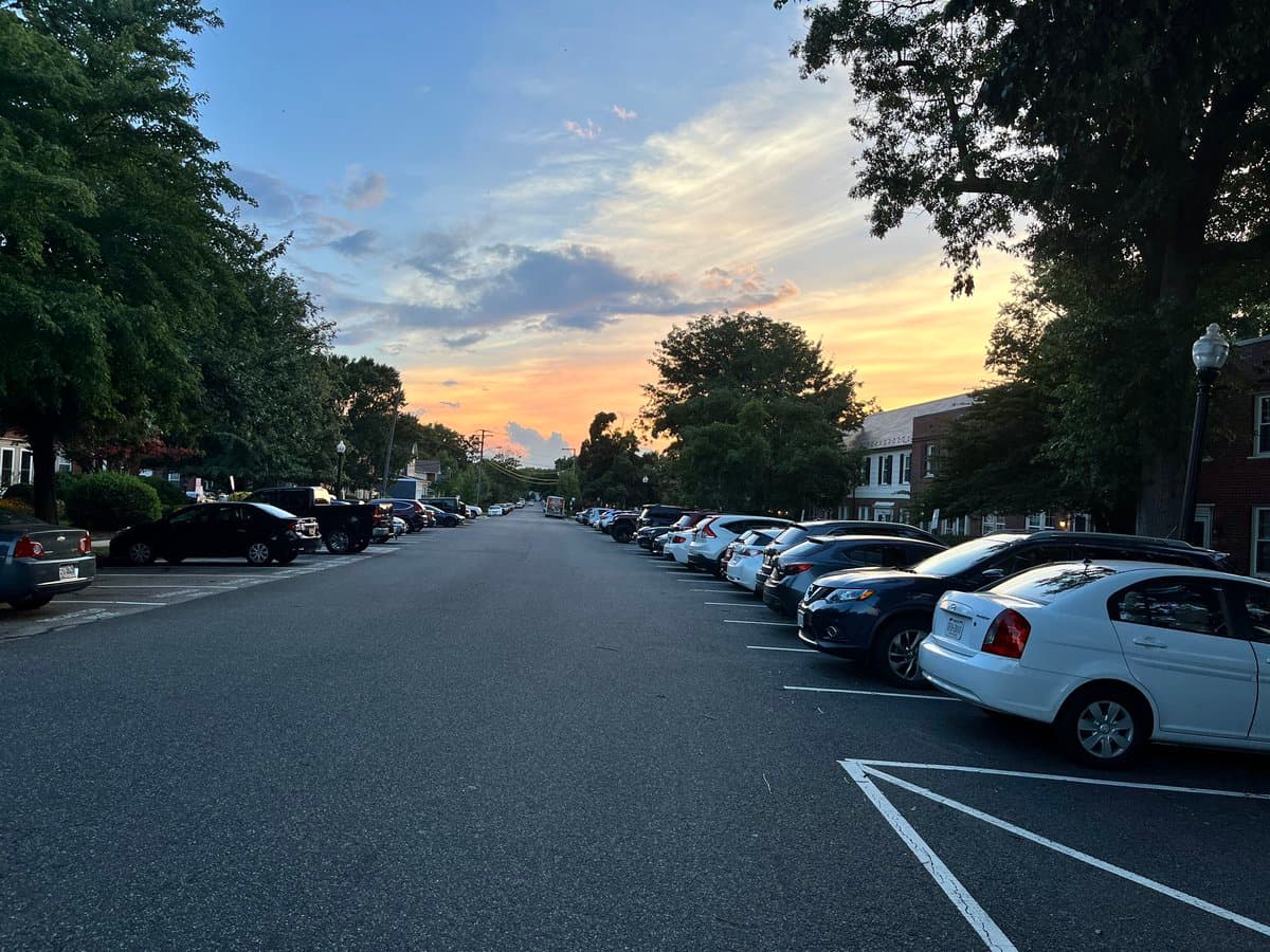 A street-level photograph taken at dusk showing a residential road lined with parked cars on both sides, flanked by trees and houses under a colorful sunset sky.
