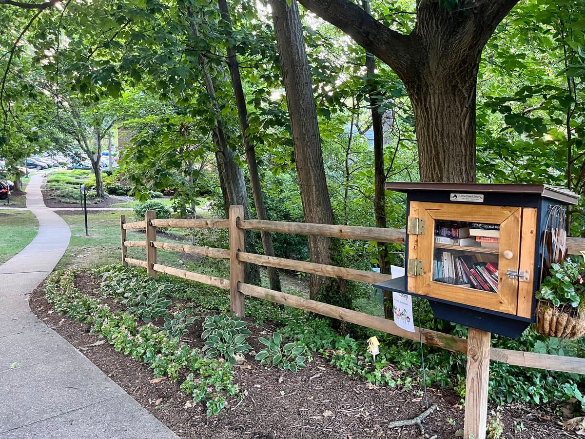 A photograph of a small, wooden Little Free Library box mounted on a wooden fence in a landscaped garden area next to a paved sidewalk. The scene is set in a wooded area with trees and greenery.