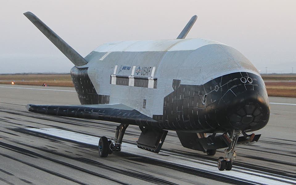 A photograph of the X-37B spaceplane on a runway, featuring its black heat shield and white body with visible markings.