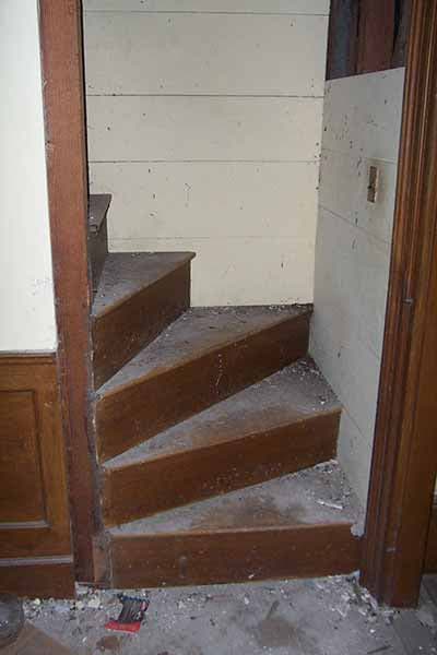 A photograph showing a narrow, steep set of wooden winder stairs in an older home, with visible dust and debris on the steps and floor. The staircase is framed by wooden door trim and white paneled walls.
