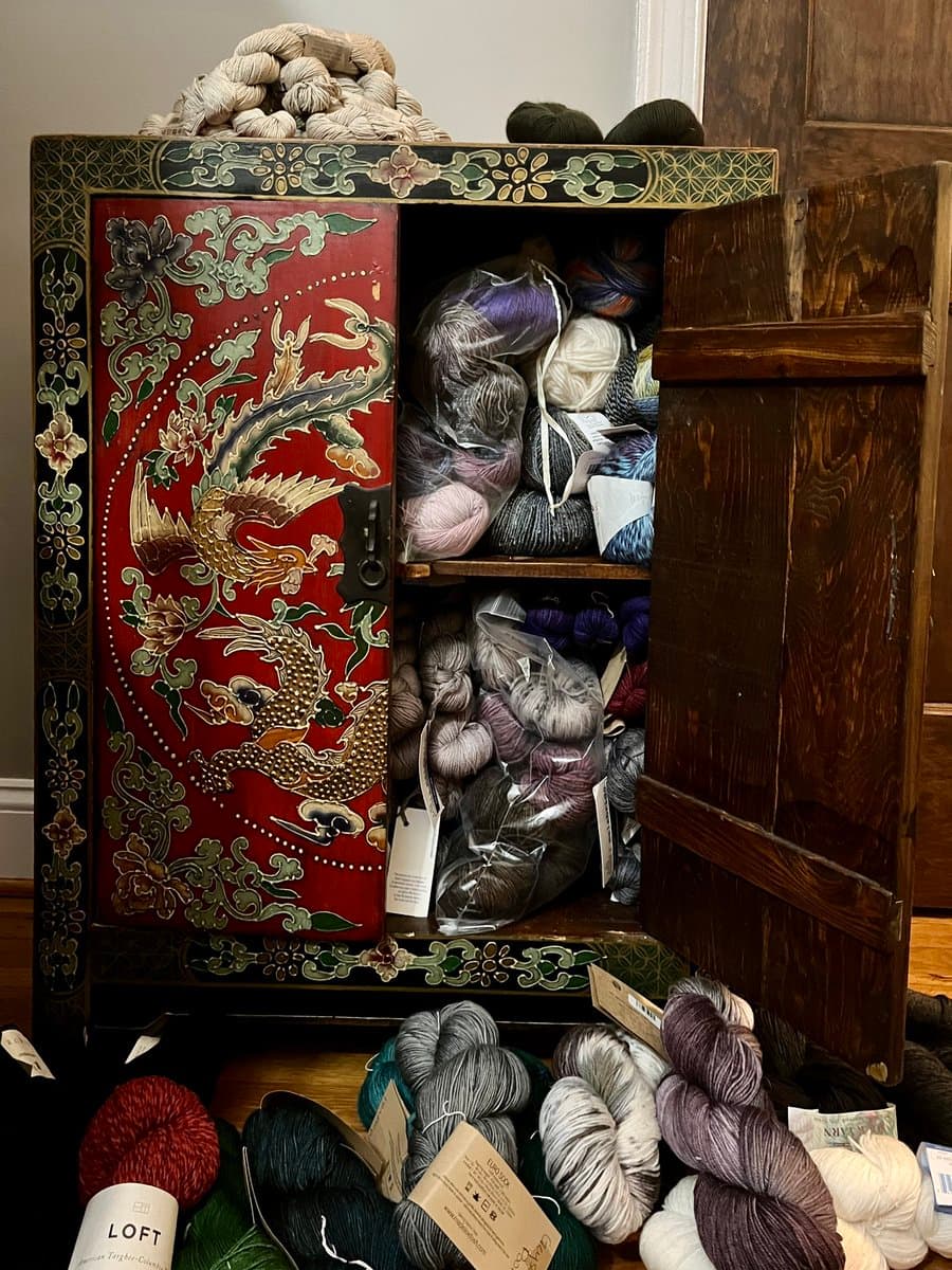 A photograph of an open, ornate red and gold lacquered cabinet filled with skeins of yarn, with more yarn piled on top and on the floor in front of it.