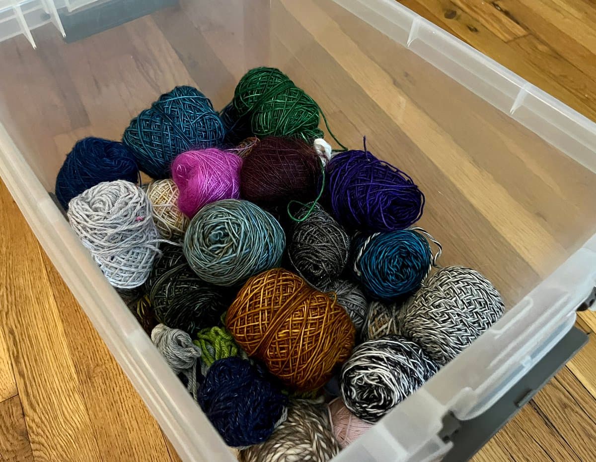 A clear plastic storage bin filled with numerous balls of yarn in various colors, including blue, green, purple, pink, and brown, sitting on a wooden floor.