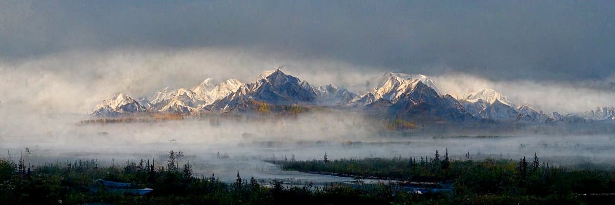 A wide landscape photograph showing a snow-capped mountain range partially obscured by low-hanging clouds and fog, with a misty valley and sparse trees in the foreground.