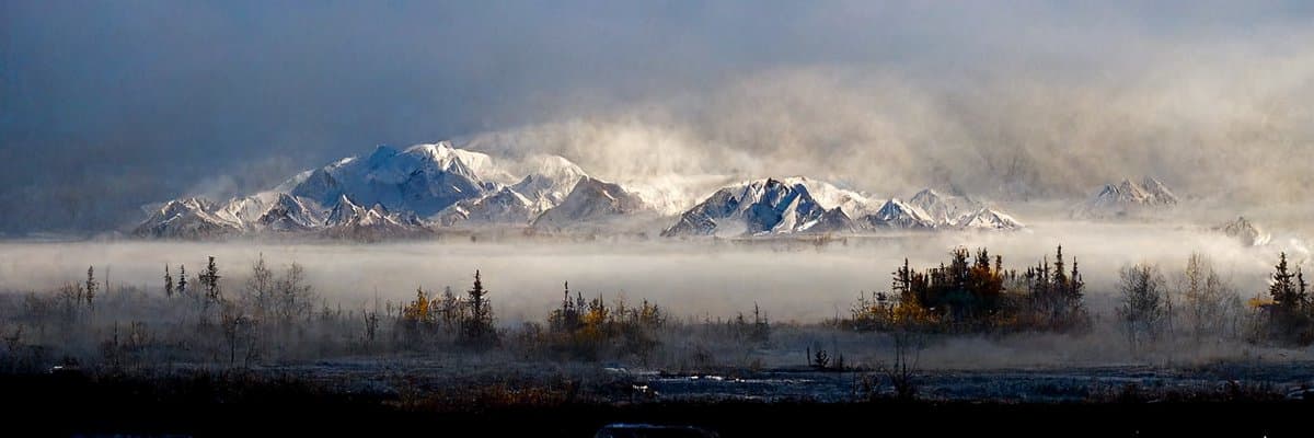 A panoramic landscape photograph showing a snow-capped mountain range partially obscured by low-lying clouds and fog, with a foreground of misty tundra and sparse trees.