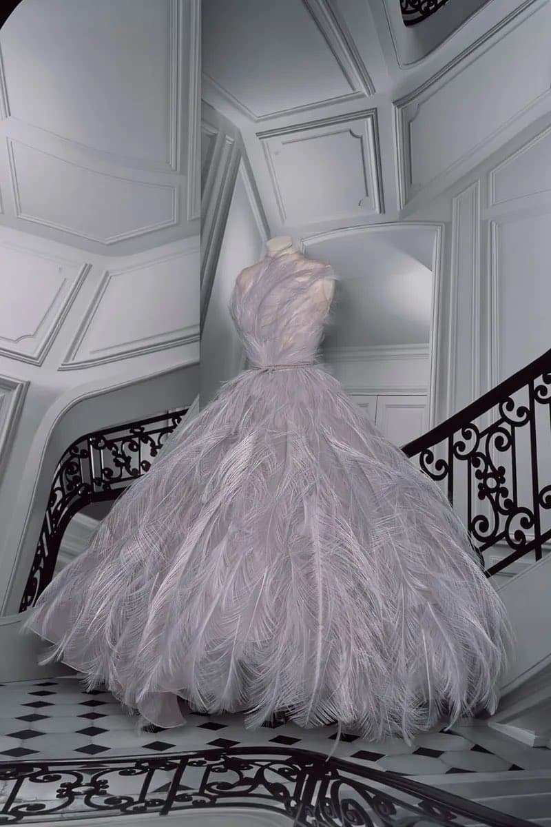 A large, voluminous white gown made of feathers is displayed on a mannequin in an ornate, all-white interior with a black-and-white checkered floor and decorative black iron railings.