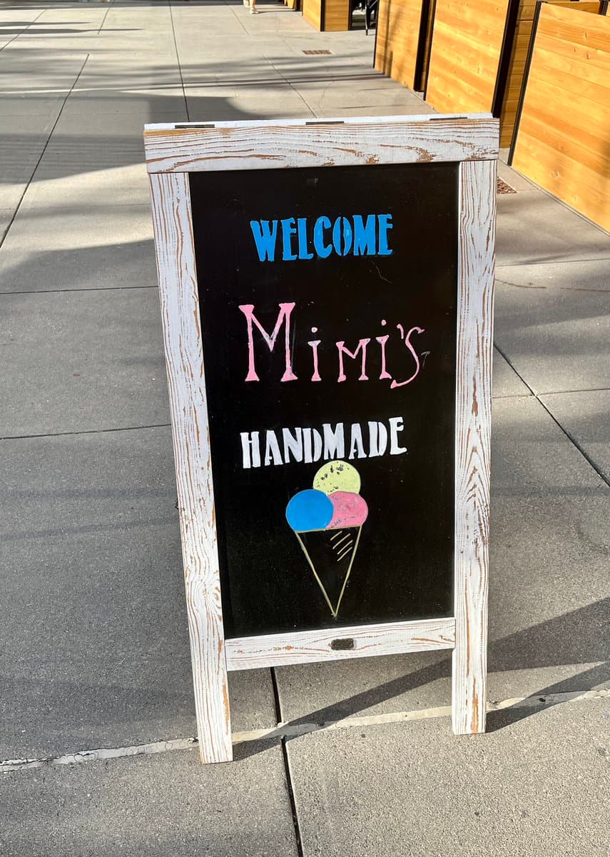 A white wooden A-frame chalkboard sign standing on a sidewalk, featuring hand-painted text and a colorful illustration of an ice cream cone. The sign advertises a business called Mimi's Handmade.