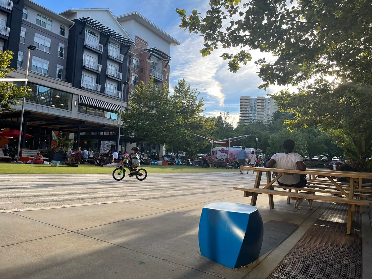 A wide-angle outdoor photograph of a public plaza featuring a child riding a small bicycle on a paved area, with a Hello Kitty-themed food truck parked in the background near a grassy lawn and apartment buildings.