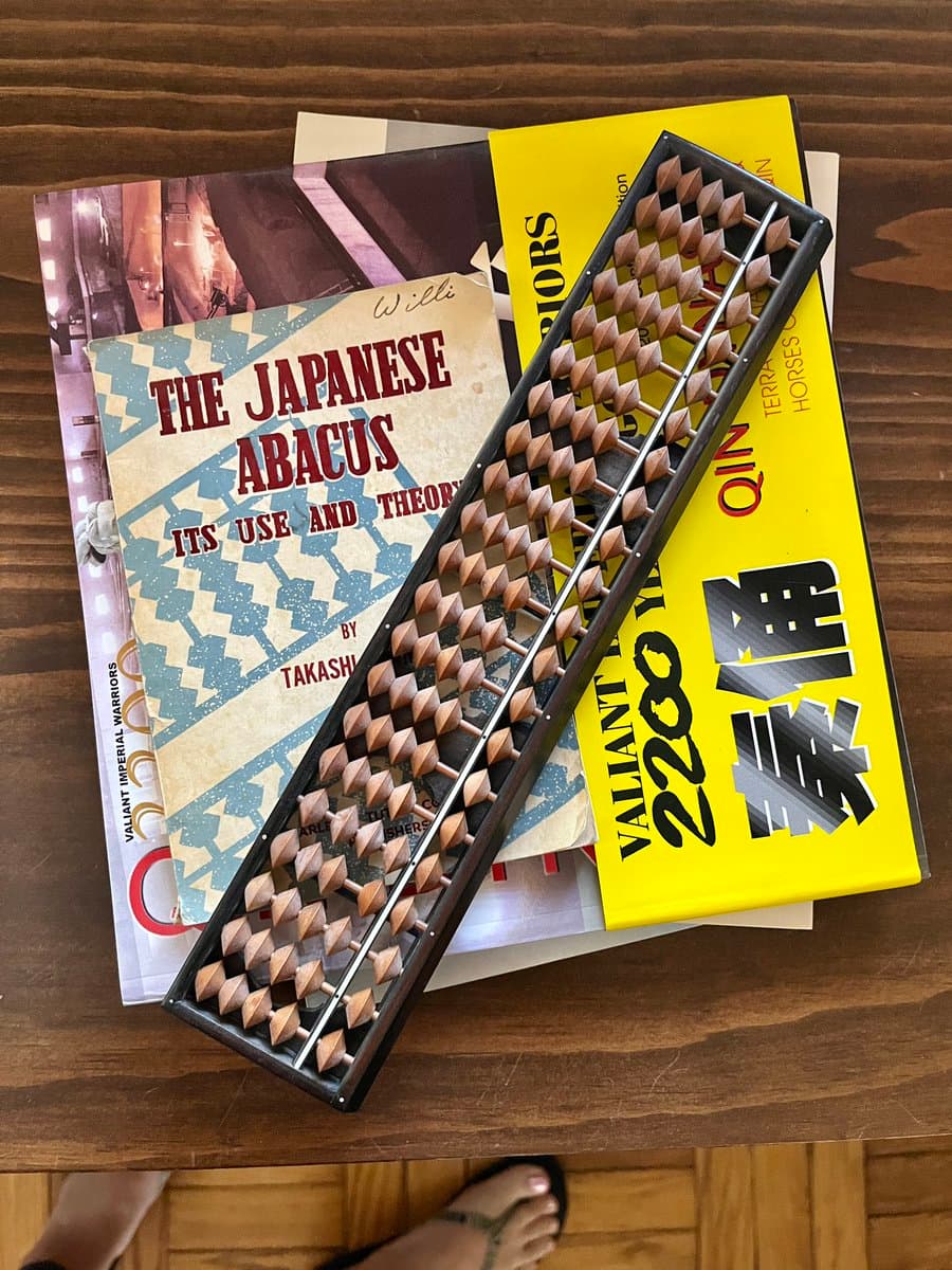 A photograph of a wooden abacus resting on a stack of books, including a manual titled 'The Japanese Abacus' and a yellow box for a 'Valiant 2200' game.
