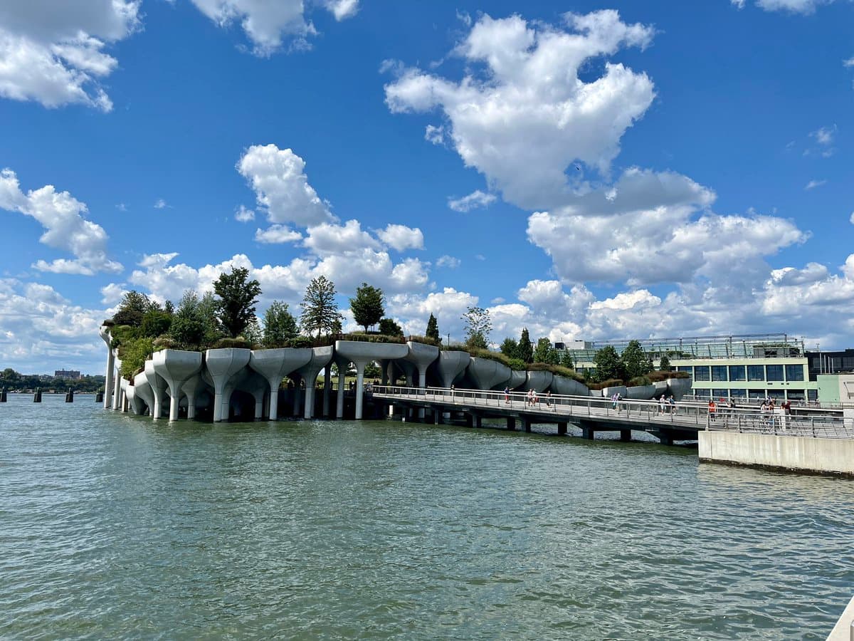 A wide-angle photograph of the High Line's elevated park structure extending over a body of water on a sunny day with a blue, partly cloudy sky. The concrete, bowl-shaped planters are filled with trees and shrubs, and a pedestrian walkway runs alongside the structure with several people visible on it.