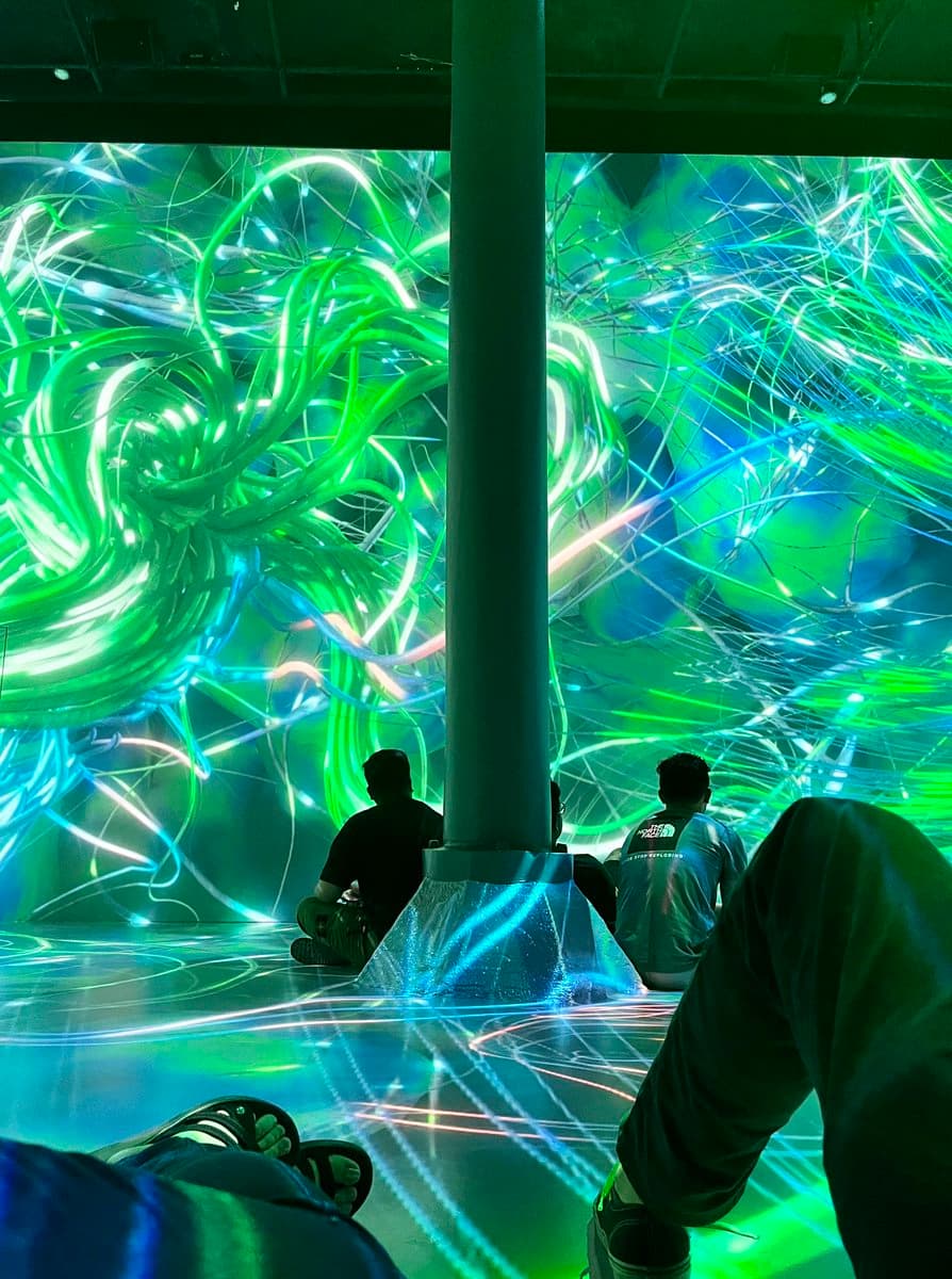 A first-person view of an immersive digital art installation featuring swirling, neon-green and blue light patterns projected onto the walls and floor. Several people are sitting on the reflective floor in front of a central pillar, observing the dynamic light display.