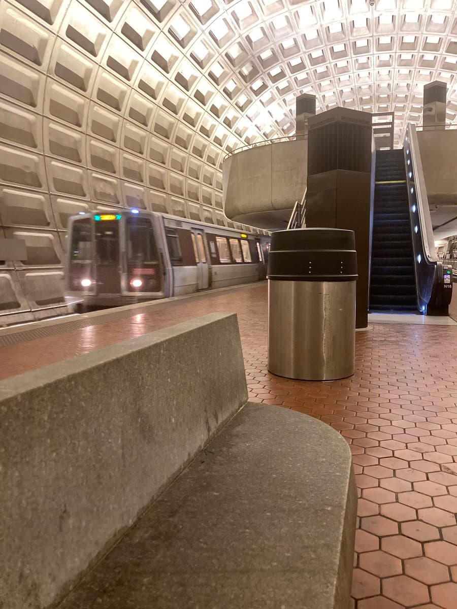 A photograph of a Washington D.C. Metro station interior, featuring a train arriving at the platform, a curved concrete wall, and a distinctive coffered ceiling.