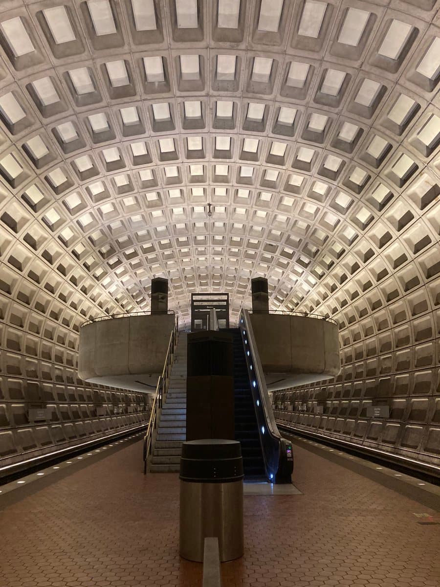 A symmetrical, low-angle photograph of a Washington D.C. Metro station interior, showcasing its distinctive coffered concrete ceiling and a central escalator leading to an upper level.