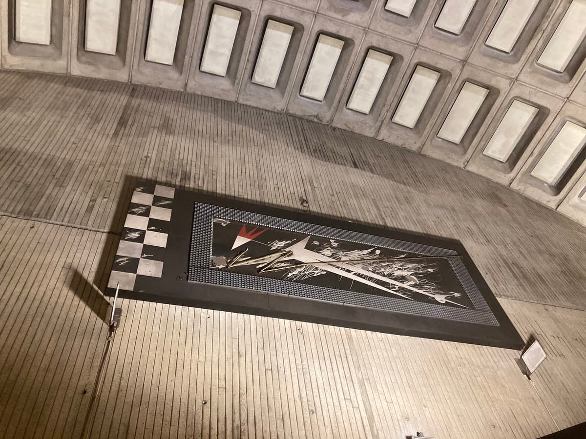 A low-angle photograph of a Washington D.C. Metro station interior, featuring a large, rectangular mosaic artwork embedded in the concrete floor. The image highlights the station's brutalist architecture, characterized by a curved wall with a grid of recessed, illuminated rectangular panels.