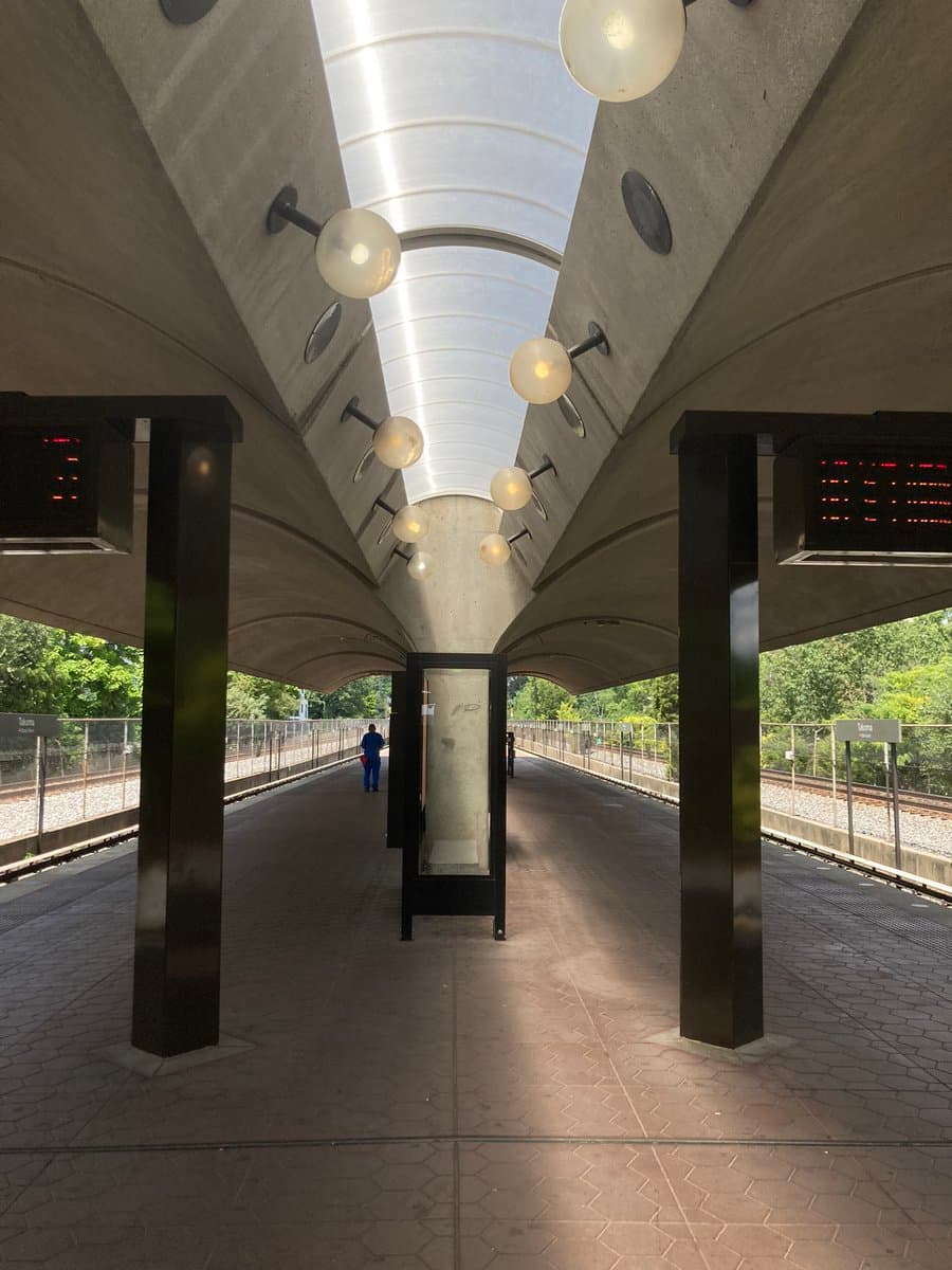 A photograph of a modern, open-air metro station platform featuring a distinctive curved concrete roof with a central skylight and spherical light fixtures. The view looks down the length of the platform, which is flanked by train tracks and greenery, with a few people visible in the distance.
