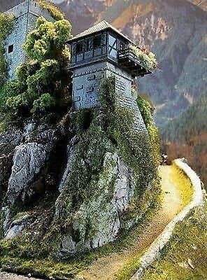 A photograph of a stone watchtower built on a rocky cliffside, surrounded by greenery and mountains. The structure features a wooden upper level with railings and is situated next to a winding dirt path.