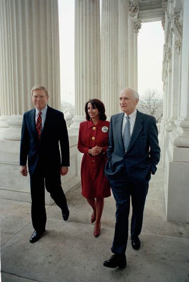 A photograph of three people walking on a stone walkway in front of a building with large white columns. The man on the left wears a dark suit and striped tie, the woman in the center wears a red dress, and the man on the right wears a blue suit and patterned tie.
