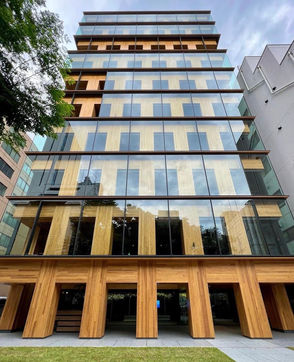 A low-angle exterior photograph of a modern multi-story building featuring a prominent facade of light-colored wood paneling and large glass windows. The structure is supported by thick, angled wooden columns at the ground level, set against a cloudy sky and adjacent to other urban buildings.