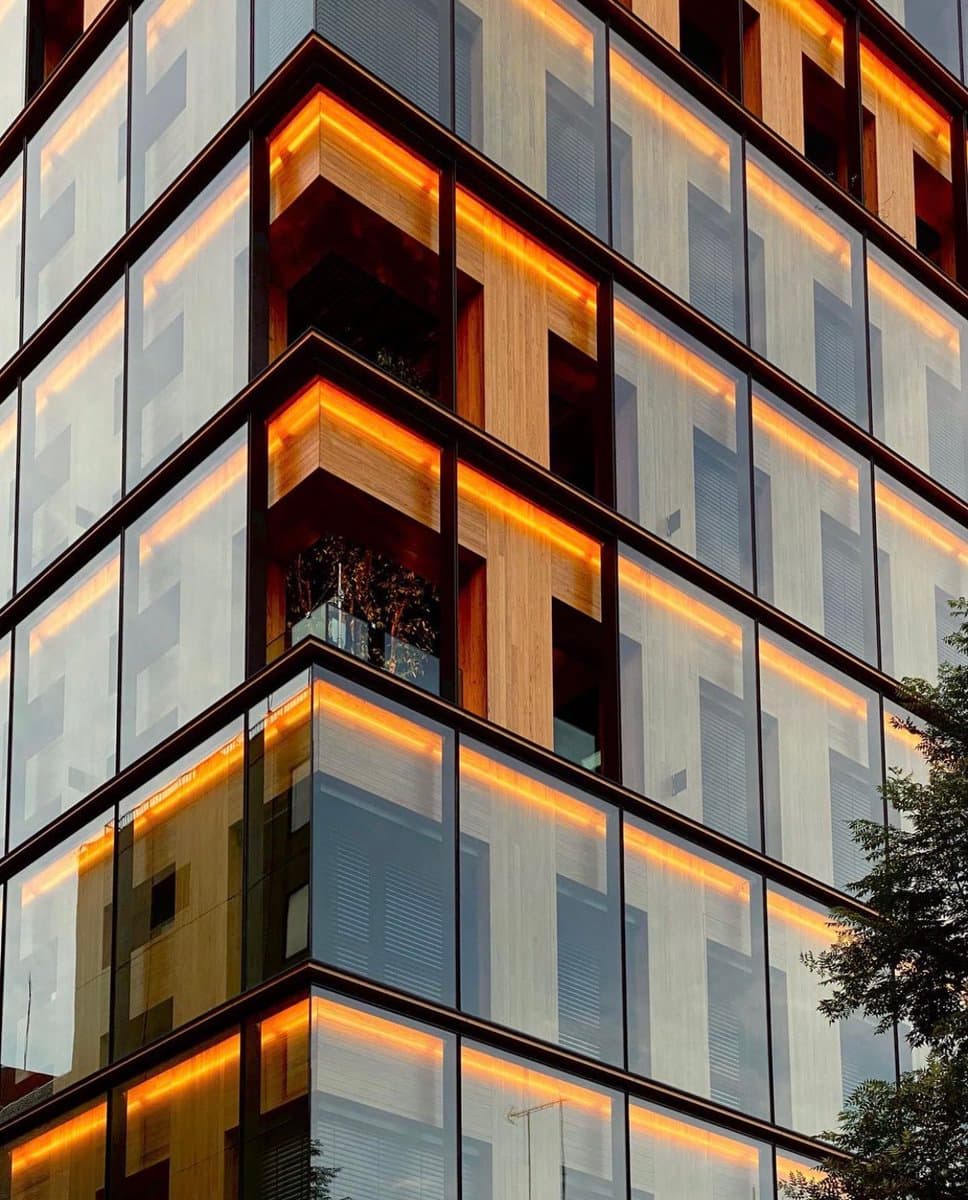 A low-angle architectural photograph of a modern high-rise building featuring a grid of glass windows and wooden paneling. Warm, orange accent lighting is installed along the top of each floor's interior, highlighting the structure's geometric design.
