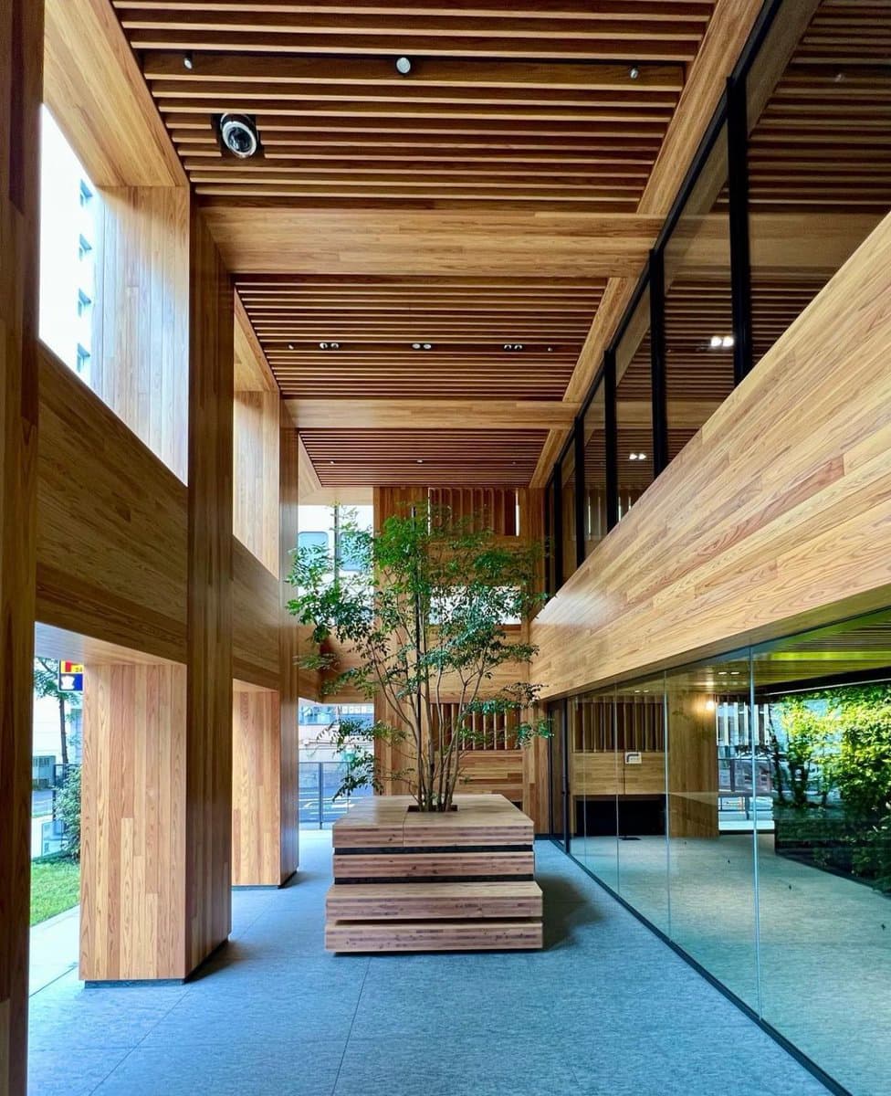 A photograph of a modern interior space featuring extensive use of light-colored wood paneling on the walls, ceiling, and a central planter box. The room has a multi-level design with glass railings and large windows, showcasing a contemporary architectural style with natural materials.