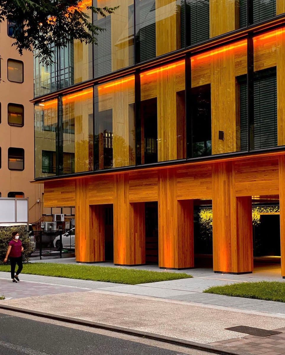 A street-level photograph of a modern multi-story building featuring a prominent wood-paneled facade with large glass windows and illuminated horizontal accents. A person wearing a face mask walks on the sidewalk in the foreground, with a grassy strip separating the walkway from the building's wooden columns.