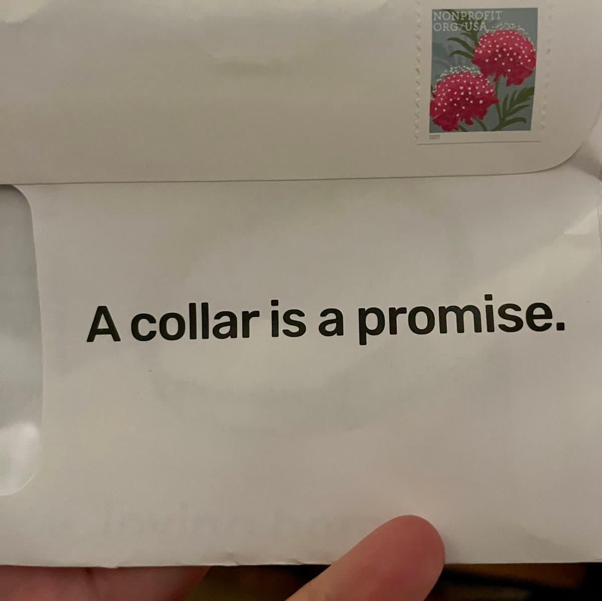 A close-up photo of a white envelope held by a hand, featuring a printed message and a postage stamp. The stamp depicts two pink flowers and is labeled 'NONPROFIT ORG-USA'.