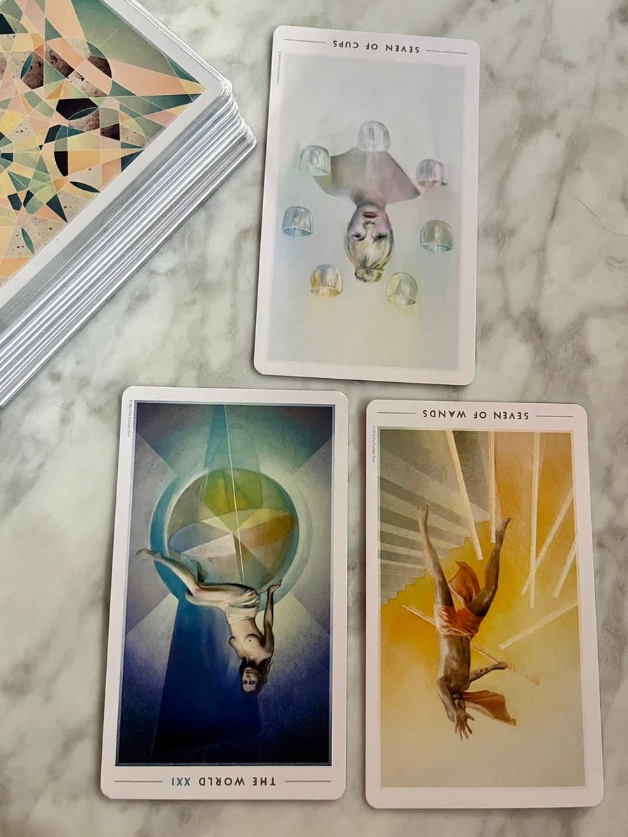 A photograph of three reversed tarot cards laid out on a white marble surface next to a stack of cards. The cards are titled "SEVEN OF CUPS", "THE WORLD XXI", and "SEVEN OF WANDS".
