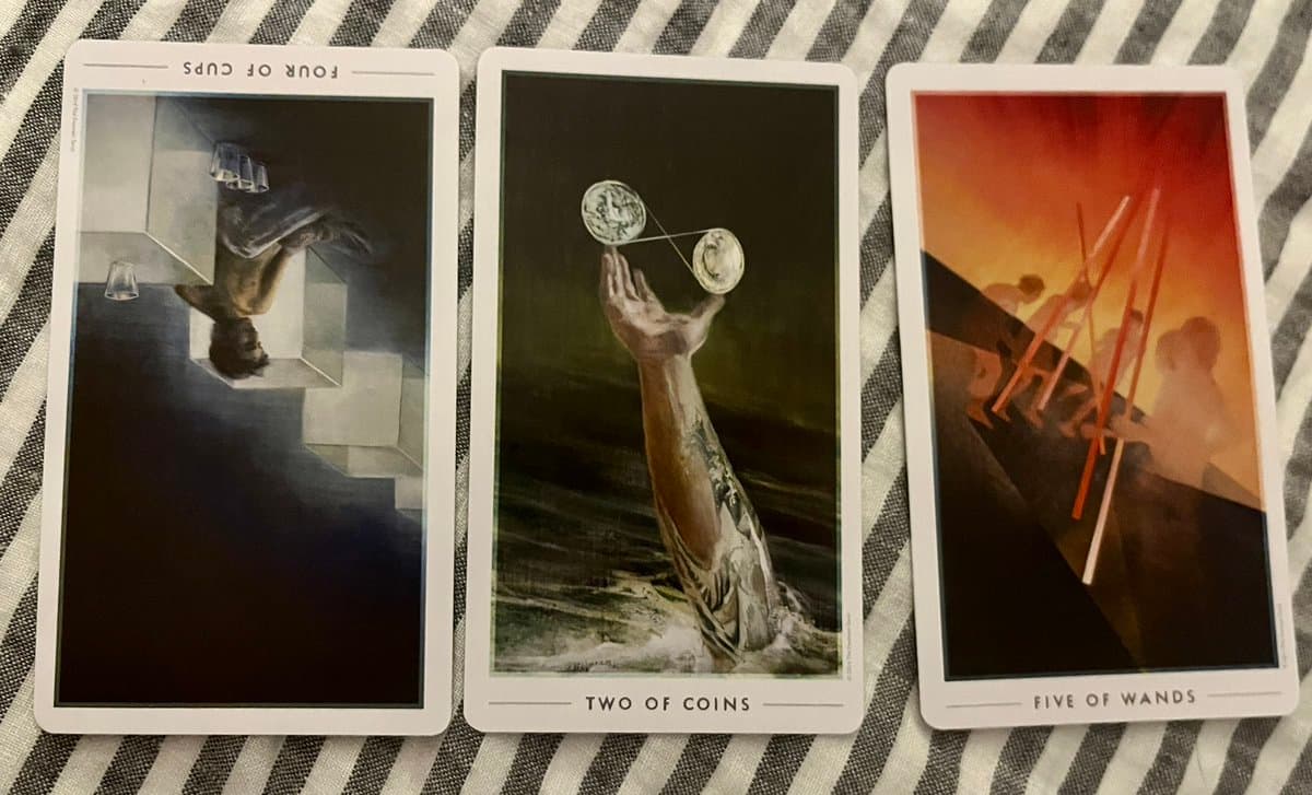 Three tarot cards are laid out on a striped fabric surface. The left card is the Four of Cups (upside down), the middle card is the Two of Coins, and the right card is the Five of Wands.