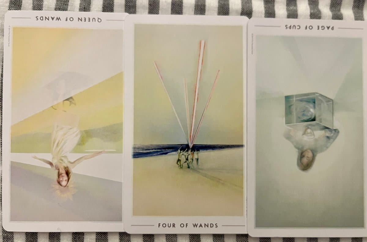 A photograph of three tarot cards laid out on a striped fabric surface. The cards are the Queen of Wands (upright), Four of Wands (upright), and Page of Cups (upside down).