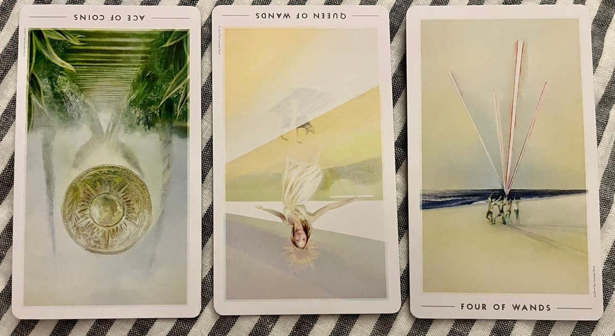 A photograph of three tarot cards laid out on a black and white striped cloth. The cards are the Ace of Coins (upright), Queen of Wands (reversed), and Four of Wands (upright).