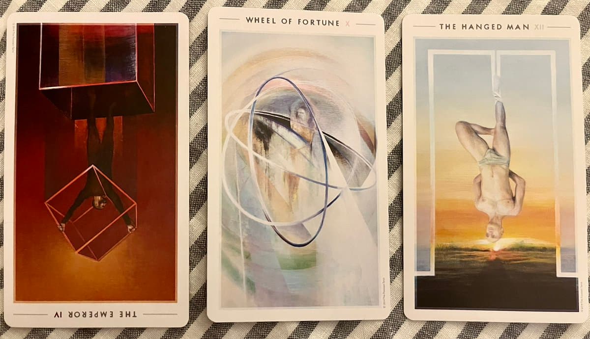 A photograph of three tarot cards laid out on a striped fabric surface. The cards are The Emperor (reversed), The Wheel of Fortune, and The Hanged Man.