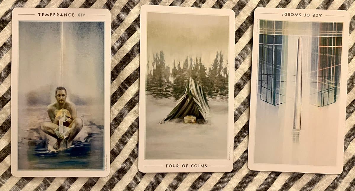 A photograph of three tarot cards laid out on a striped fabric surface. The cards are titled Temperance, Four of Coins, and Ace of Swords (reversed).