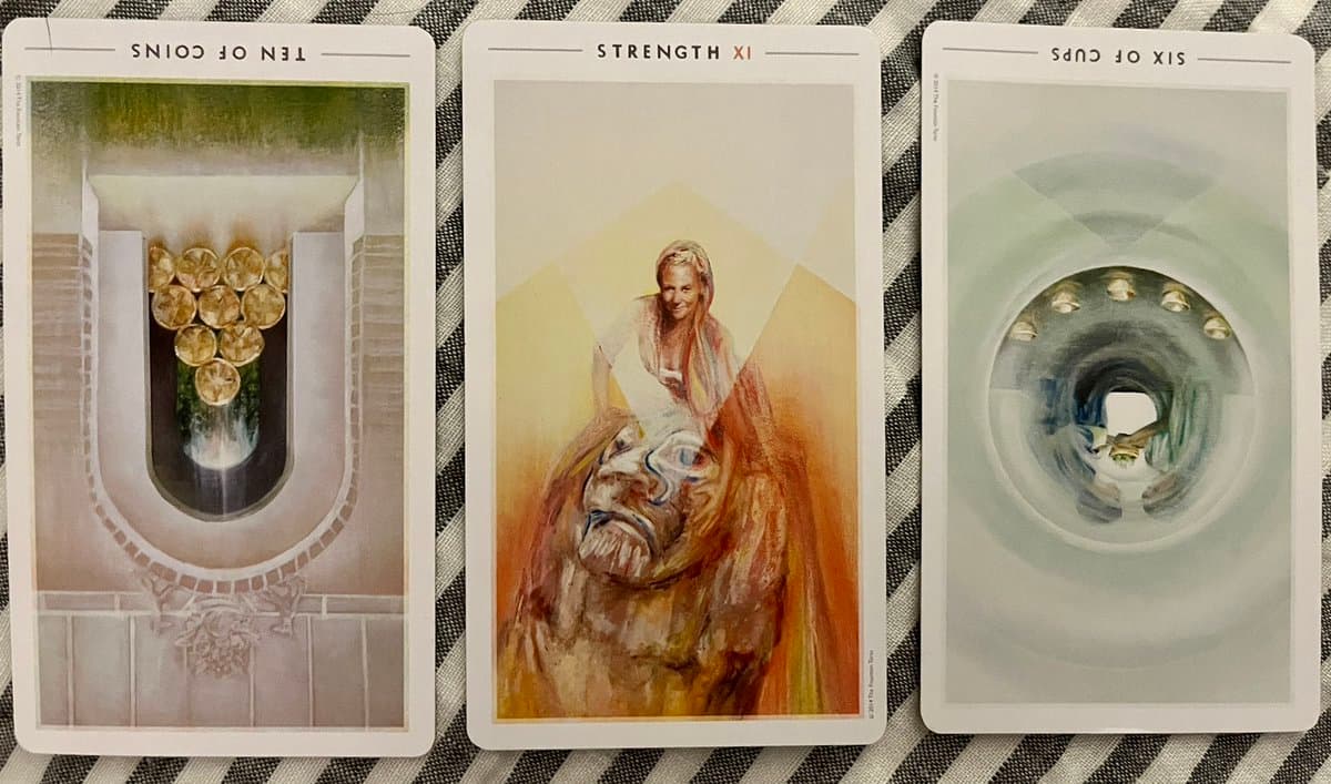 A photograph of three tarot cards laid out on a black and white striped cloth. The cards are displayed upside down, showing the Ten of Coins, Strength, and Six of Cups.