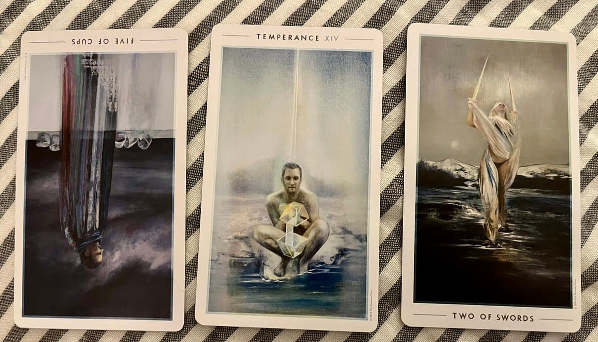 A photograph of three tarot cards laid out on a striped fabric surface. The cards are the Five of Cups (upside down), Temperance, and the Two of Swords.