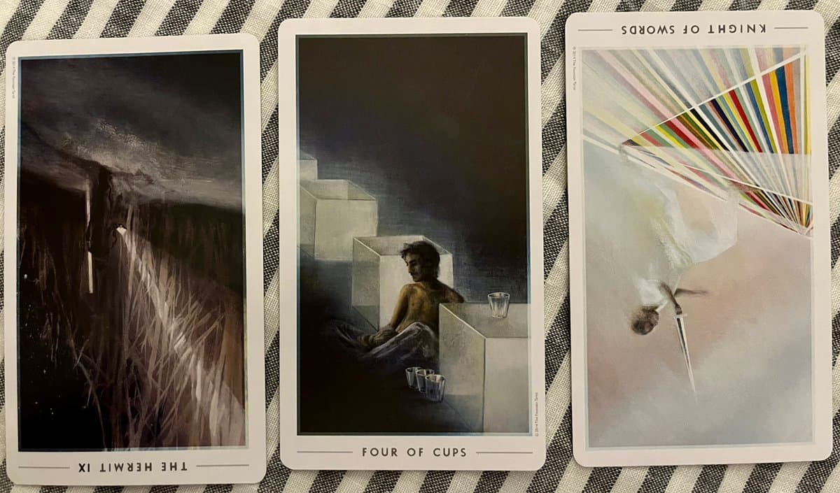 A photograph of three tarot cards laid out on a black-and-white striped fabric, showing the Hermit, Four of Cups, and Knight of Swords.