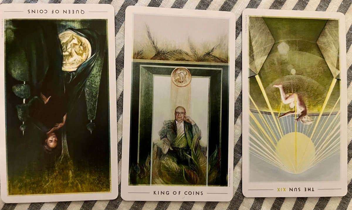 A photograph of three tarot cards laid out on a striped cloth, showing the Queen of Coins, King of Coins, and The Sun. The Queen of Coins and The Sun cards are placed upside down, indicating they are reversed.