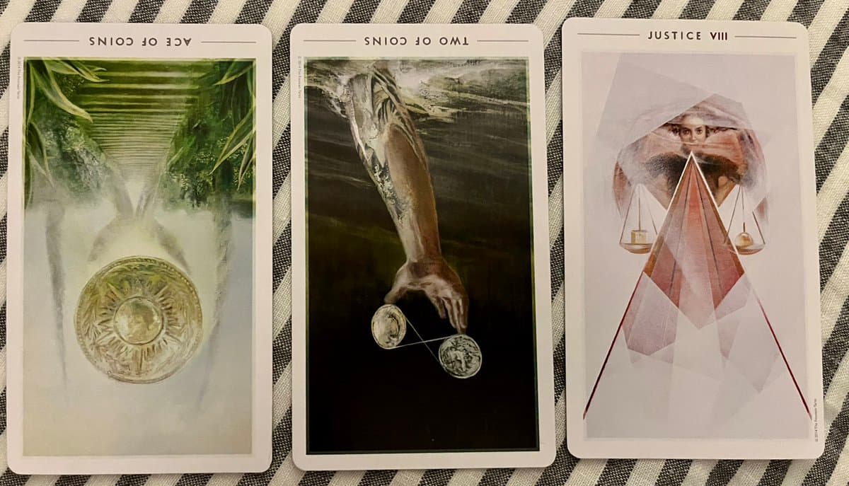 A photograph of three tarot cards laid out on a striped fabric surface, showing the Ace of Coins, Two of Coins, and Justice cards. The cards feature surreal, artistic imagery including a coin in a misty landscape, a hand holding coins, and a figure with scales.
