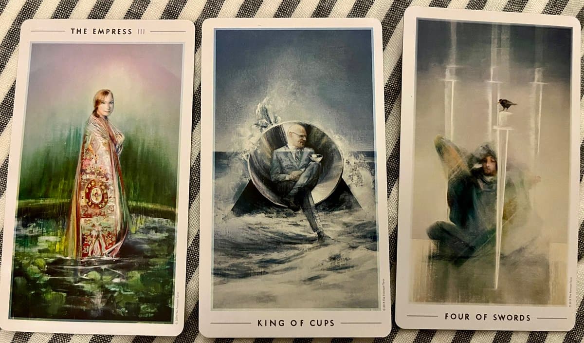A photograph of three tarot cards laid out on a striped fabric surface, showing The Empress, King of Cups, and Four of Swords.