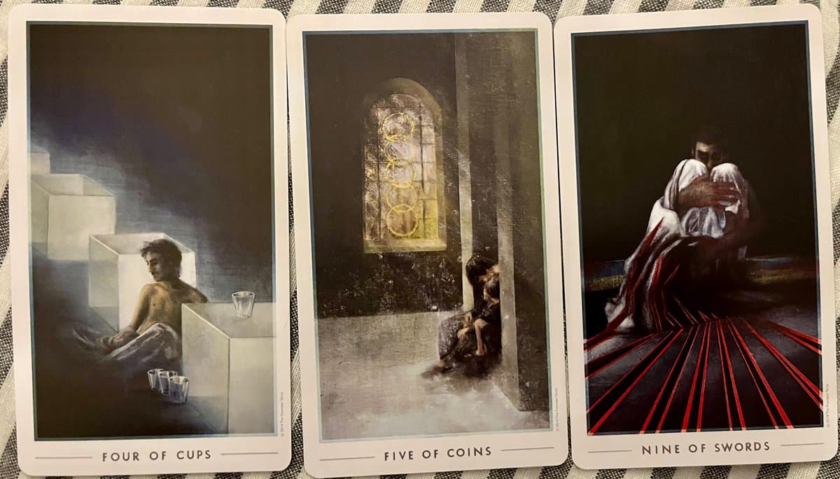 A photograph of three tarot cards laid out on a striped cloth, showing the Four of Cups, Five of Coins, and Nine of Swords.
