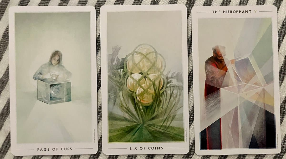 A photograph of three tarot cards laid out on a striped fabric surface. The cards, from left to right, are the Page of Cups, Six of Coins, and The Hierophant, each featuring distinct artwork and titles.
