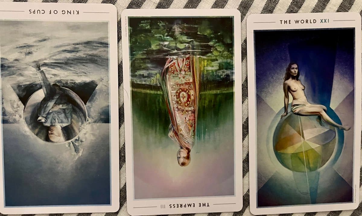 A photograph of three tarot cards laid out on a striped fabric surface. The left and center cards are placed upside down, while the right card is upright.