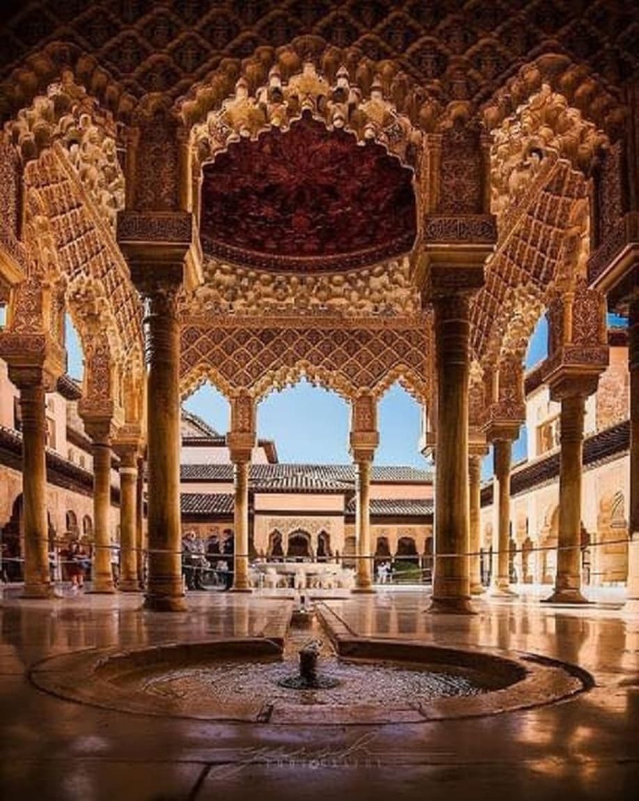 A photograph of the ornate Court of the Lions at the Alhambra in Granada, featuring intricate Moorish arches, columns, and a central fountain.
