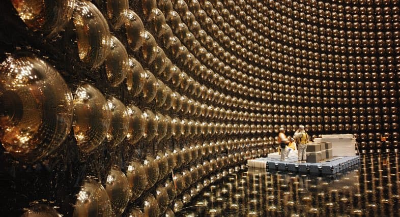 A wide-angle photograph of the interior of a massive neutrino detector, showing a curved wall of thousands of golden, spherical photomultiplier tubes. Two people in work clothes stand on a small platform in the center, providing a sense of the detector's enormous scale.
