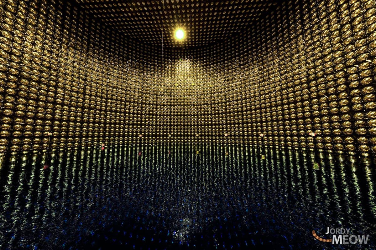 A wide-angle photograph of the interior of a neutrino detector, showing a vast, curved wall of golden photomultiplier tubes surrounding a dark, reflective pool of water. A single bright light source is visible near the top center, and a watermark is present in the bottom right corner.