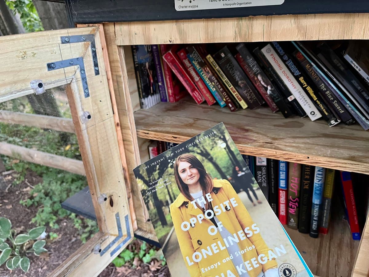 A close-up photograph of a wooden 'free book' box filled with various paperbacks, with a copy of 'The Opposite of Loneliness' by Lena Keegan held up in the foreground. The box is situated outdoors near a wooden fence and greenery.
