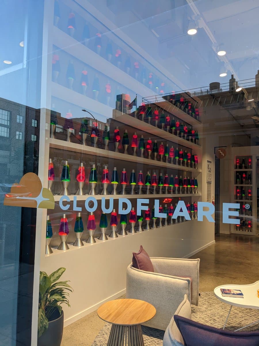 A photograph of a modern office interior featuring a large wall display of numerous colorful lava lamps arranged on shelves. The word "CLOUDFLARE" is written in large, light blue letters across the center of the display, with a logo to the left.