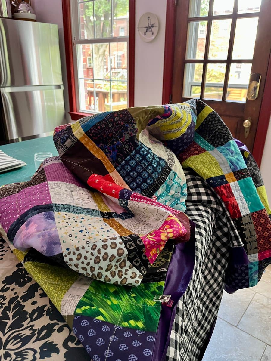 A colorful patchwork quilt made of various patterned fabric squares is draped over a chair in a room with a window and a stainless steel refrigerator in the background.