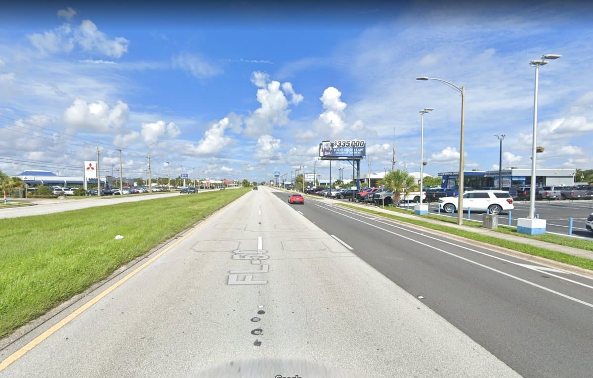 A wide-angle street-level view of a multi-lane highway with a large grassy median, flanked by commercial car dealerships and parking lots under a blue sky with scattered clouds.