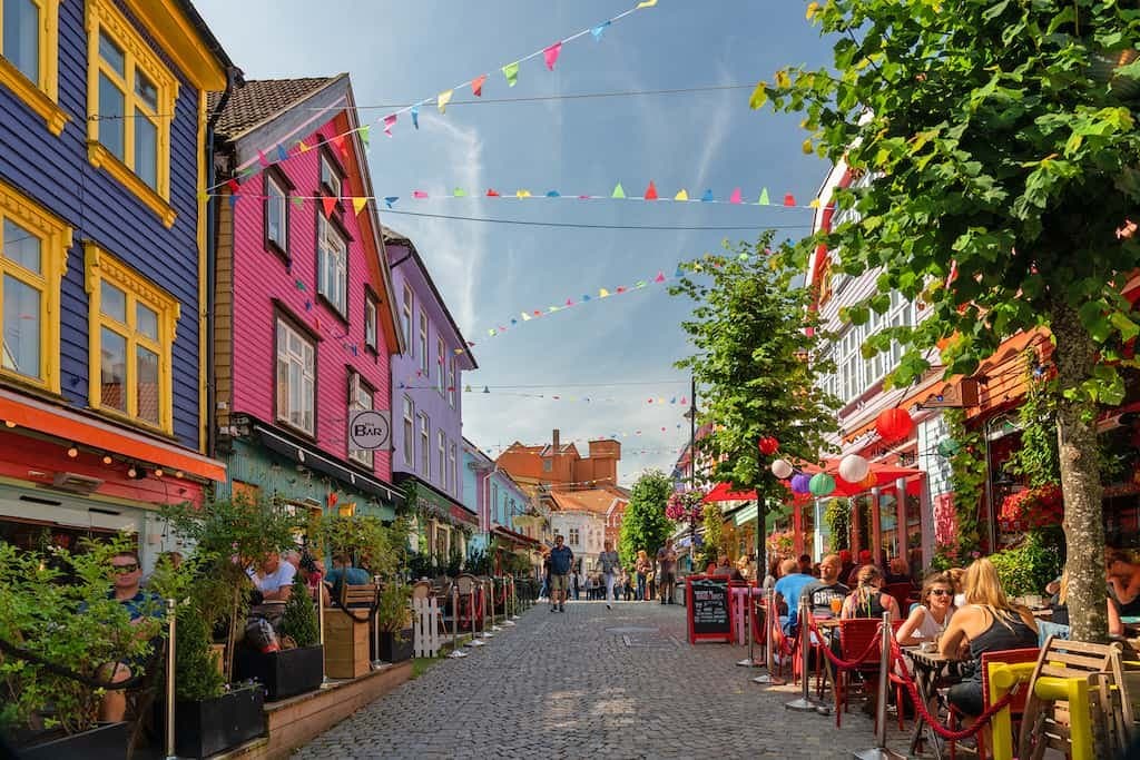 A vibrant, pedestrian-only street lined with colorful buildings in shades of blue, pink, and purple. The cobblestone path is decorated with strings of multicolored pennant flags overhead, and people are seated at outdoor cafe tables on both sides.