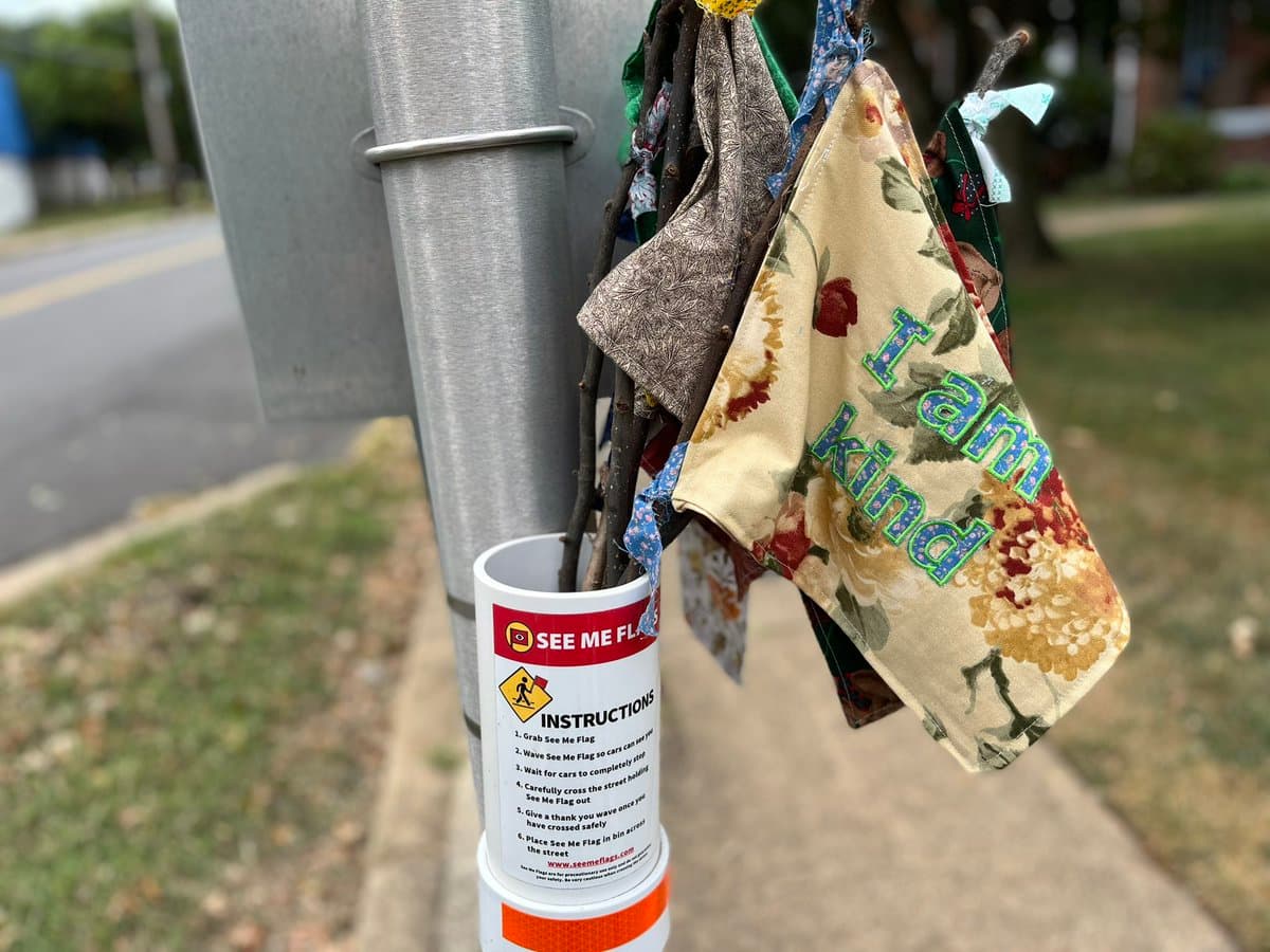 A close-up photograph of a metal pole on a sidewalk with several small, handmade fabric flags tied to it. One prominent flag features a floral pattern with the words "I am kind" in green and blue lettering. Below the flags, a white cylindrical container labeled "SEE ME FLAG" holds the flag poles.
