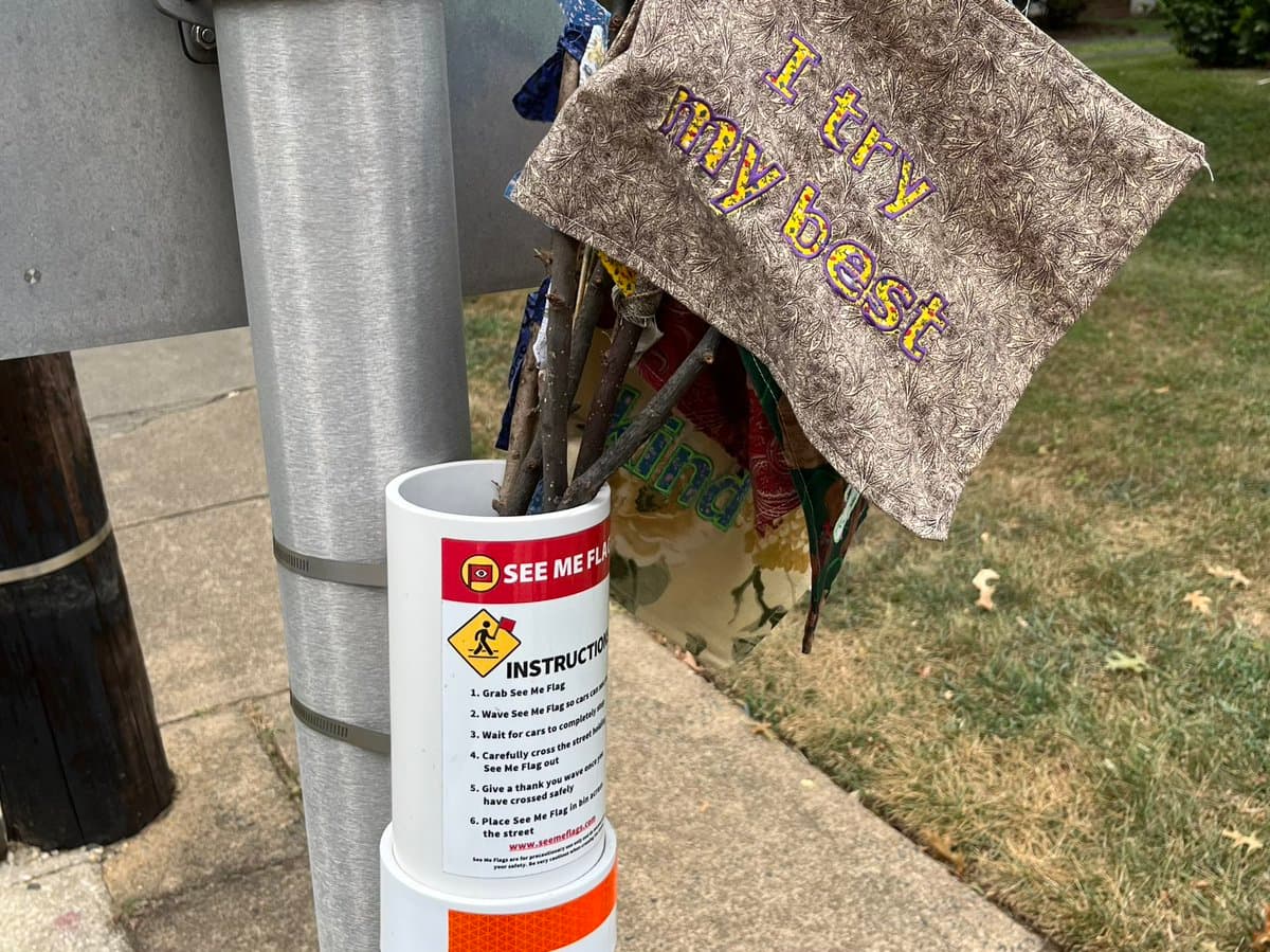 A close-up photograph of a white traffic cone on a sidewalk holding a bundle of sticks and handmade flags, including one that reads "I try my best." The cone is labeled with a "SEE ME FLAG" sign and instructions for pedestrians to use the flags to signal to drivers.